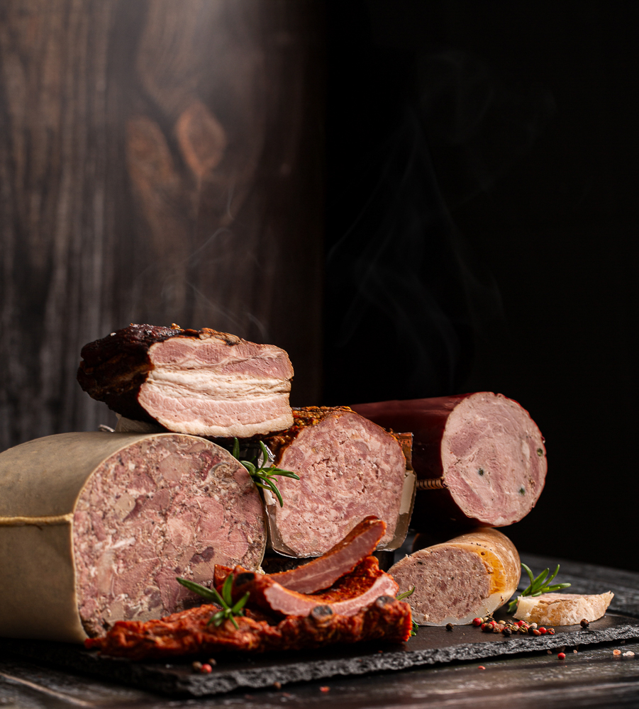 Klimatyczna fotografia produktów spożywczych – wędliny i boczki na ciemnym, drewnianym tle.